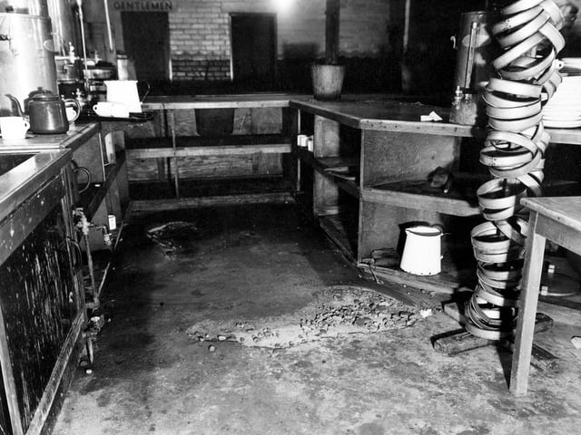 Inside the British Restaurant at Sheepscar. A stand of plate stackers is on the right. Wooden shelves, the oven, a tea pot, a bucket and the door to the men's toilets are visible.