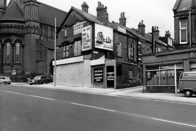 April 1980. A wholesale clothing warehouse on Roundhay Road. The front is boarded up and there are advertising hoardings on the upper floor. St. Aidan's Church is on the left with Elford Place leading off.