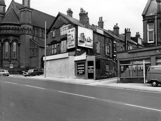 April 1980. A wholesale clothing warehouse on Roundhay Road. The front is boarded up and there are advertising hoardings on the upper floor. St. Aidan's Church is on the left with Elford Place leading off.