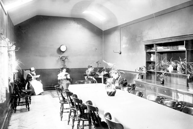 This view shows the nurses room at Seacroft Hospital, with table in centre, chairs around it. A dresser stands against the wall, nurses can be seen reading and working.