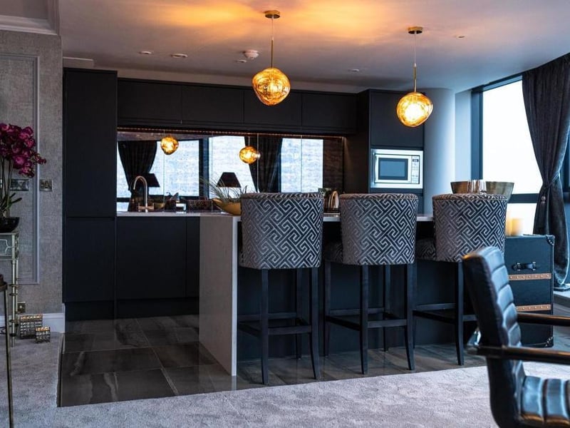 The kitchen is decorated with tiled floor, black cabinets, a mirror wall, low hanging lights and an island with high stools.