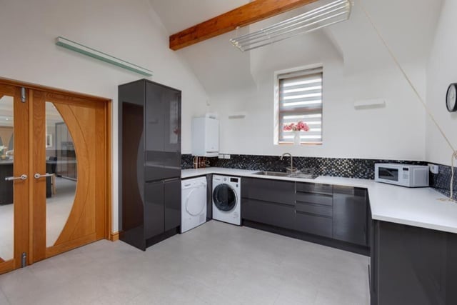 The sleek modern look of the kitchen extends into the spacious utility room which has a high ceiling with exposed beams. Fitted wall and base units have matching worksurfaces and there are tiled splashbacks.