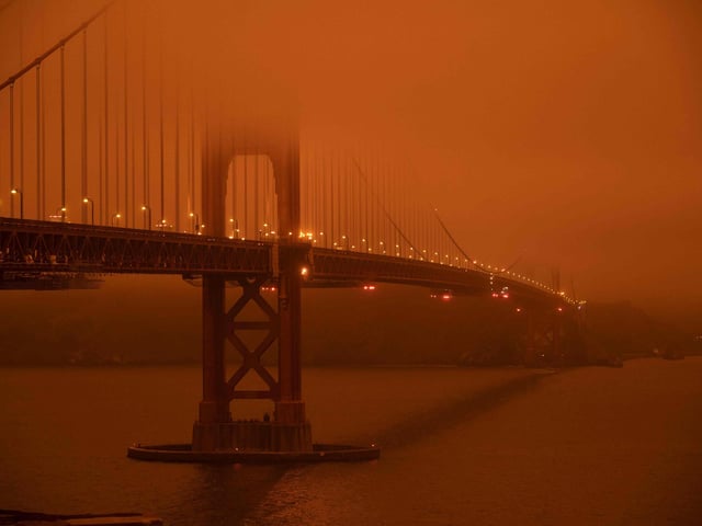 Cars drive along the San Francisco Bay Bridge under an orange smoke filled sky at midday in San Francisco.