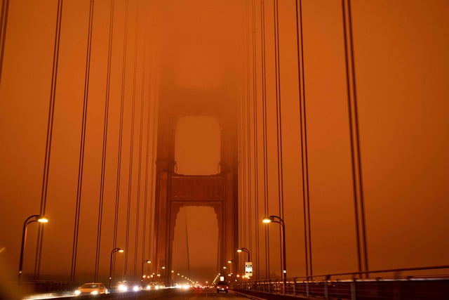 Cars drive along the San Francisco Bay Bridge under an orange smoke filled sky at midday.