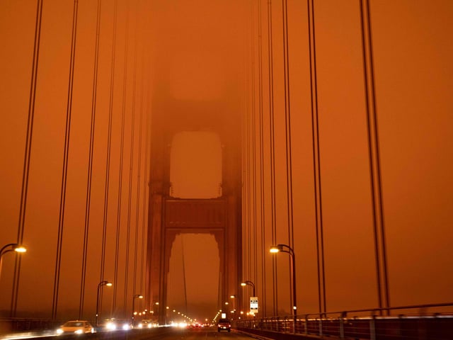 Cars drive along the San Francisco Bay Bridge under an orange smoke filled sky at midday.