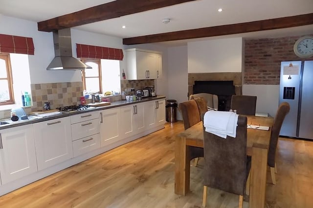 The spacious farmhouse style kitchen has exposed ceiling beams and a fireplace with  stone surround and stone hearth that houses an electric fire. Fitted units have round edge worktops and tiled splashbacks. Integrated appliances include refrigerator and dishwasher, double oven with cupboards above and below and inset four burner propane gas hob with stainless steel extractor hood above.