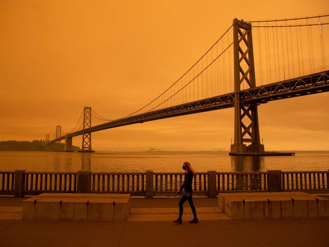 A woman walks along The Embarcadero under an orange smoke-filled sky in San Francisco.