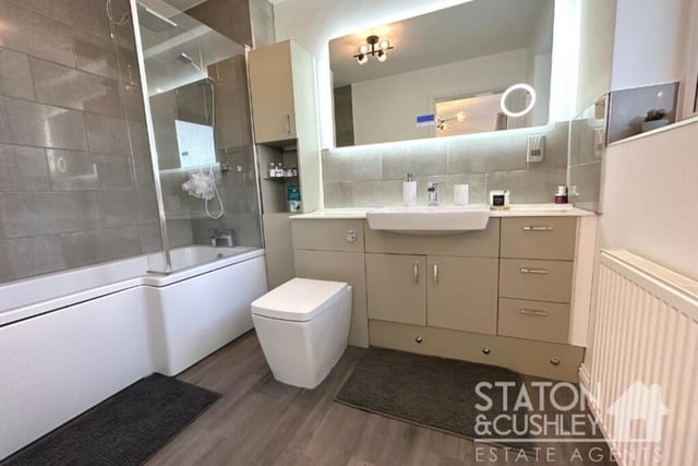 This is the en suite bathroom, accessible from the master bedroom on the ground floor. It includes a P-shaped, panelled bath with overhead mains shower, mixer tap and shower screen, as well as a vanity unit with mounted sink, low-level WC and wall-mounted mirror, plus storage.