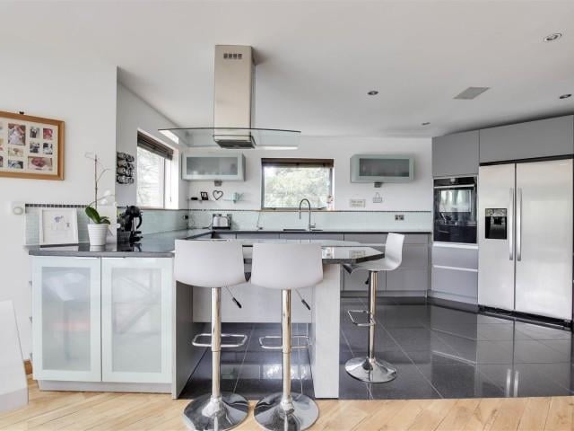 The kitchen area has a range of base and wall units with granite worktops and breakfast bar, an inverted stainless steel sink and a half with mixer taps, an integrated electric oven and grill, an electric hob with an extractor fan, space for an American style fridge freezer, under counter strip lights, tiled splash back, tiled flooring with underfloor heating, recessed spotlights and two double glazed windows.